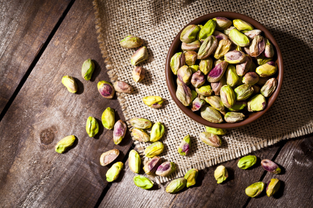 Top view of a brown bowl filled with organic pistachios shot on rustic wood table. Some almonds are out of the bowl on a burlap. Predominant colors are green and brown. DSRL studio photo taken with Canon EOS 5D Mk II and Canon EF 100mm f/2.8L Macro IS USM